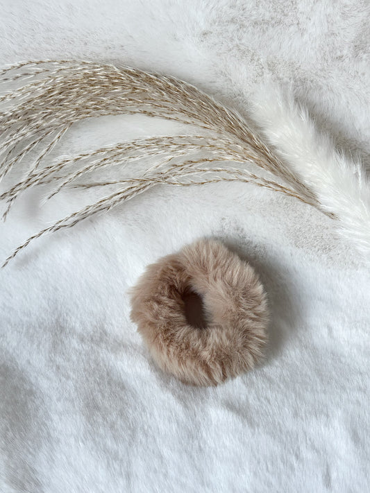 Beiger flauschiger Scrunchie aus weichem Kunstfell, ästhetisch arrangiert auf hellem Stoff mit Pampasgras – stilvolles Haar-Accessoire von Javi Jewelery
