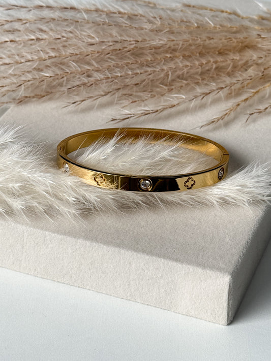 Eleganter goldener Armreif mit eingraviertem Kleeblatt-Design und funkelnden Zirkonia-Steinen, stilvoll präsentiert auf beigem Stoff mit Pampasgras – Javi Jewelery
