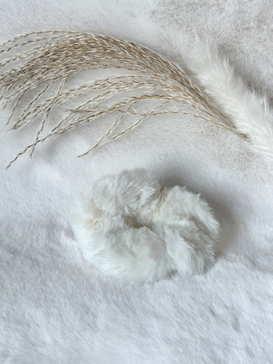 Weißer flauschiger Scrunchie aus weichem Kunstfell, stilvoll arrangiert auf hellem Stoff mit Pampasgras – elegantes Haar-Accessoire von Javi Jewelery