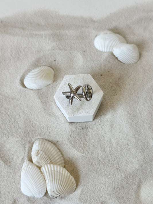 Silberner Ring mit Seestern- und Muschel-Design, kunstvoll präsentiert auf hellem Sand mit Muscheldekor – maritimer Statement-Schmuck von Javi Jewelery